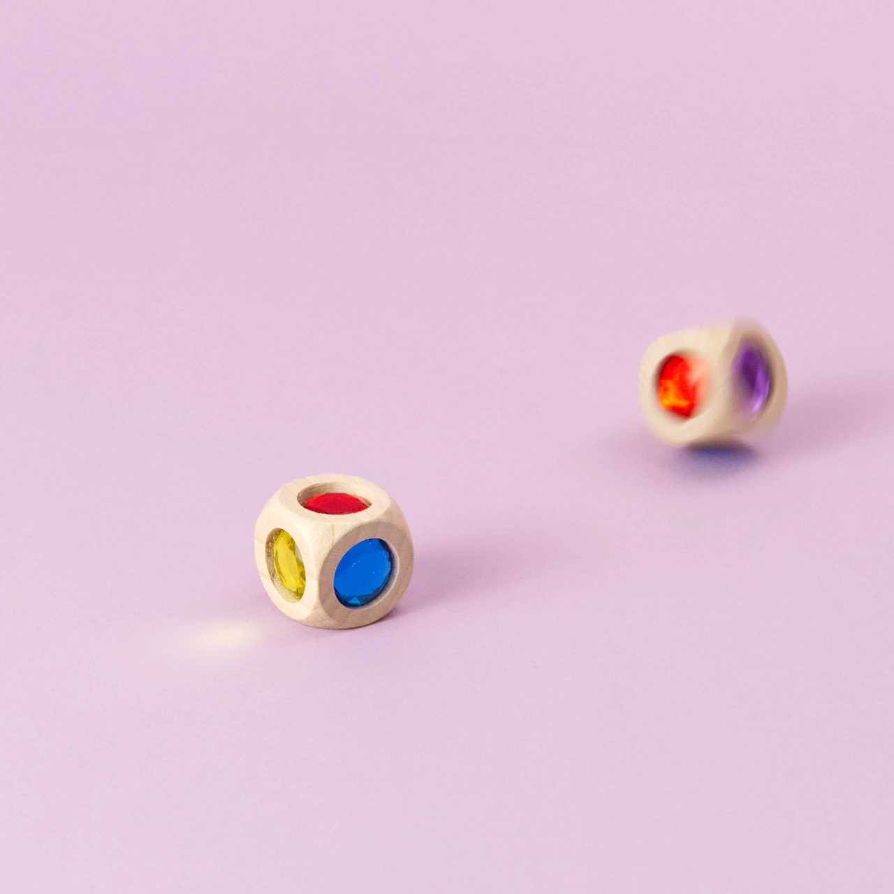 Ein Würfel aus Buchenholz mit funkelnden Acrylglas-Diamanten in Bewegung, unscharf geworfen, daneben ein liegender Würfel. Die Farben der Würfelseiten sind Rot, Blau, Grün, Gelb, Orange und Violett sichtbar.